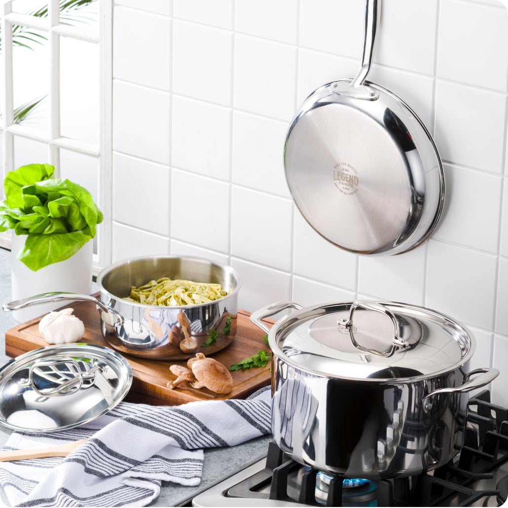 Legend Stainless fry pan hanging on a white tiled wall above a saucier of pasta and a lidded Legend stock pot on a lit gas burner
